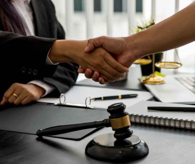 Nad biurkiem z dokumentami, notatnikiem i młotkiem sędziowskim, dłonie dwóch osób w uścisku symbolizują zawarcie ugody lub porozumienia prawnego