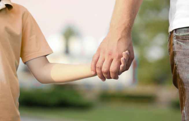 <br />
Zbliżenie na dłoń osoby dorosłej trzymającej dłoń dziecka, na niewyraźnym tle zieleni i słońca, symbolizujące opiekę i wsparcie