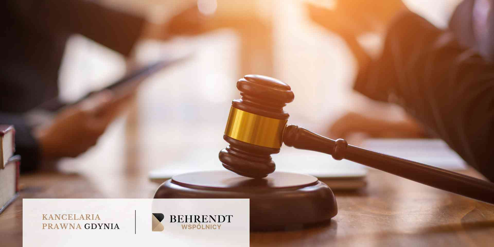 
Na drewnianym stole leży młotek sędziowski z metalowym akcentem w ciepłym, rozświetlonym wnętrzu, a w tle widać dłonie osób przeglądających dokumenty