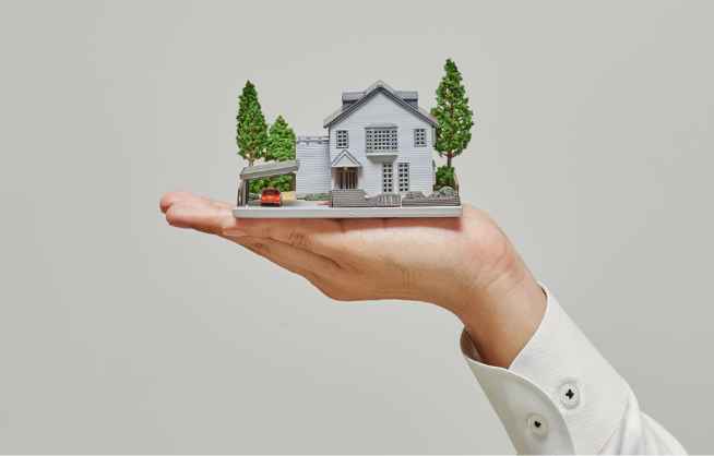 Dłoń w mankiecie białej koszuli trzyma miniaturowy model białego domu z garażem i dwoma drzewami, na jednolitym, jasnym tle.