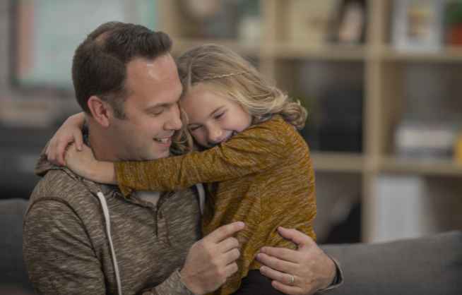 Uśmiechnięty mężczyzna siedzi na kanapie i obejmuje dziewczynkę z jasnymi włosami, która przytula się do niego.