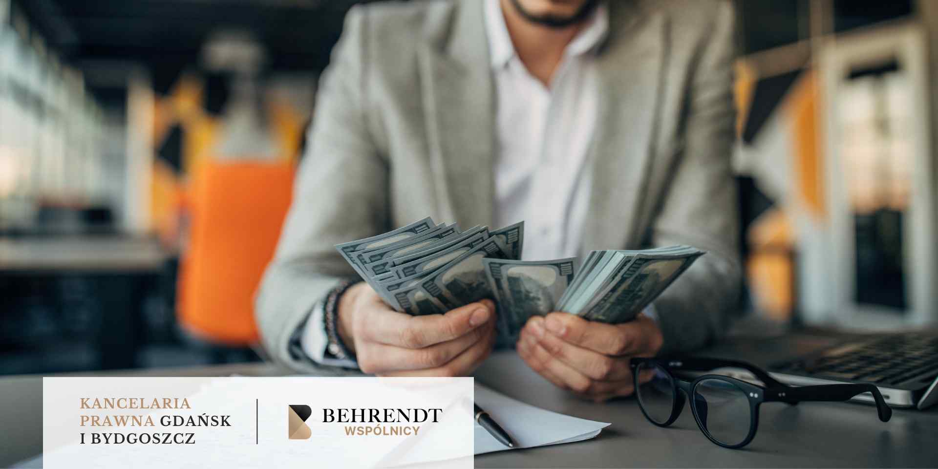 Mężczyzna w jasnej marynarce trzyma w dłoniach pliki banknotów, stojąc przy biurku z okularami