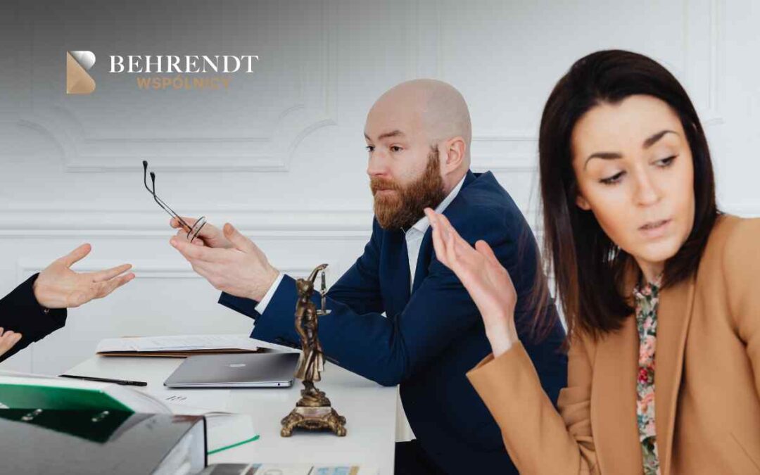 Łysy, brodaty prawnik w niebieskim garniturze i kobieta w beżowej marynarce intensywnie gestykulują podczas rozmowy przy biurku, na którym stoi figurka Temidy i laptop