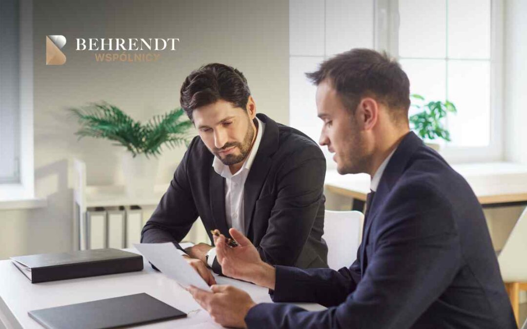 Dwóch mężczyzn w garniturach siedzi przy biurku, intensywnie analizując dokumenty w biurze