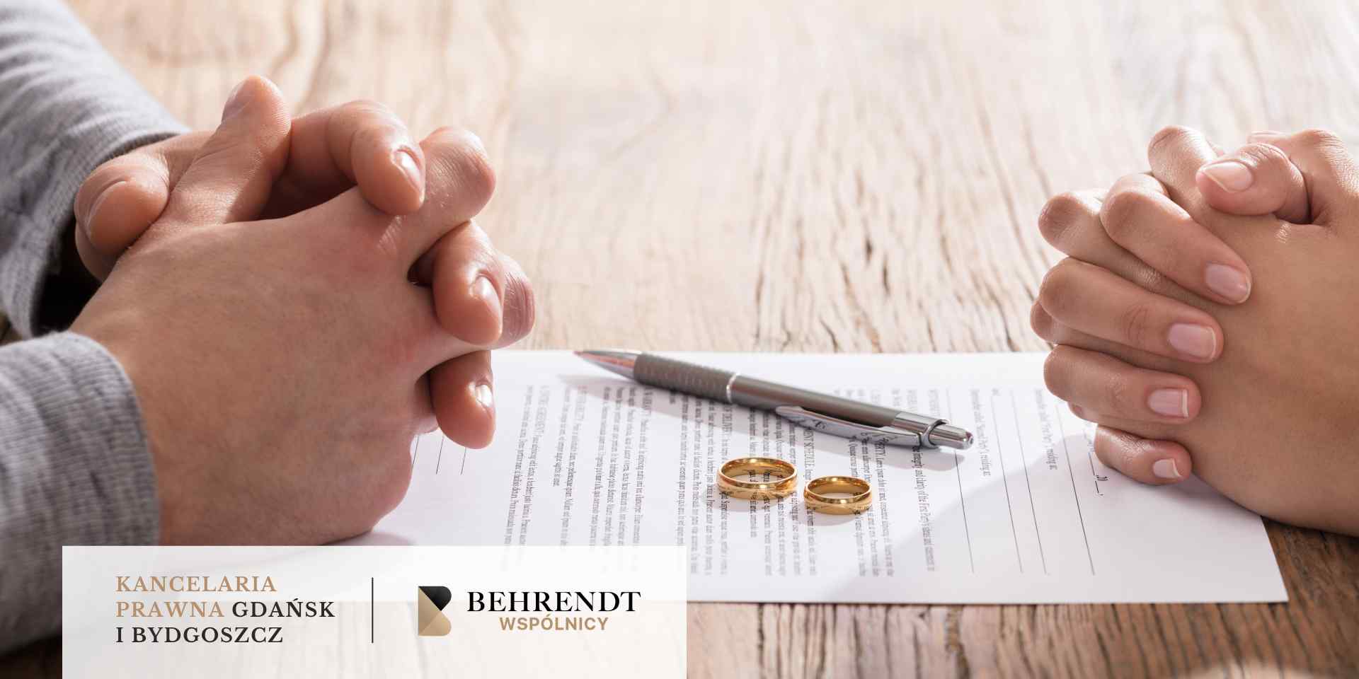 Na drewnianym stole leży dokument prawny, na którym spoczywają dwie złote obrączki i długopis, a po obu stronach widać splecione dłonie dwóch osób.
