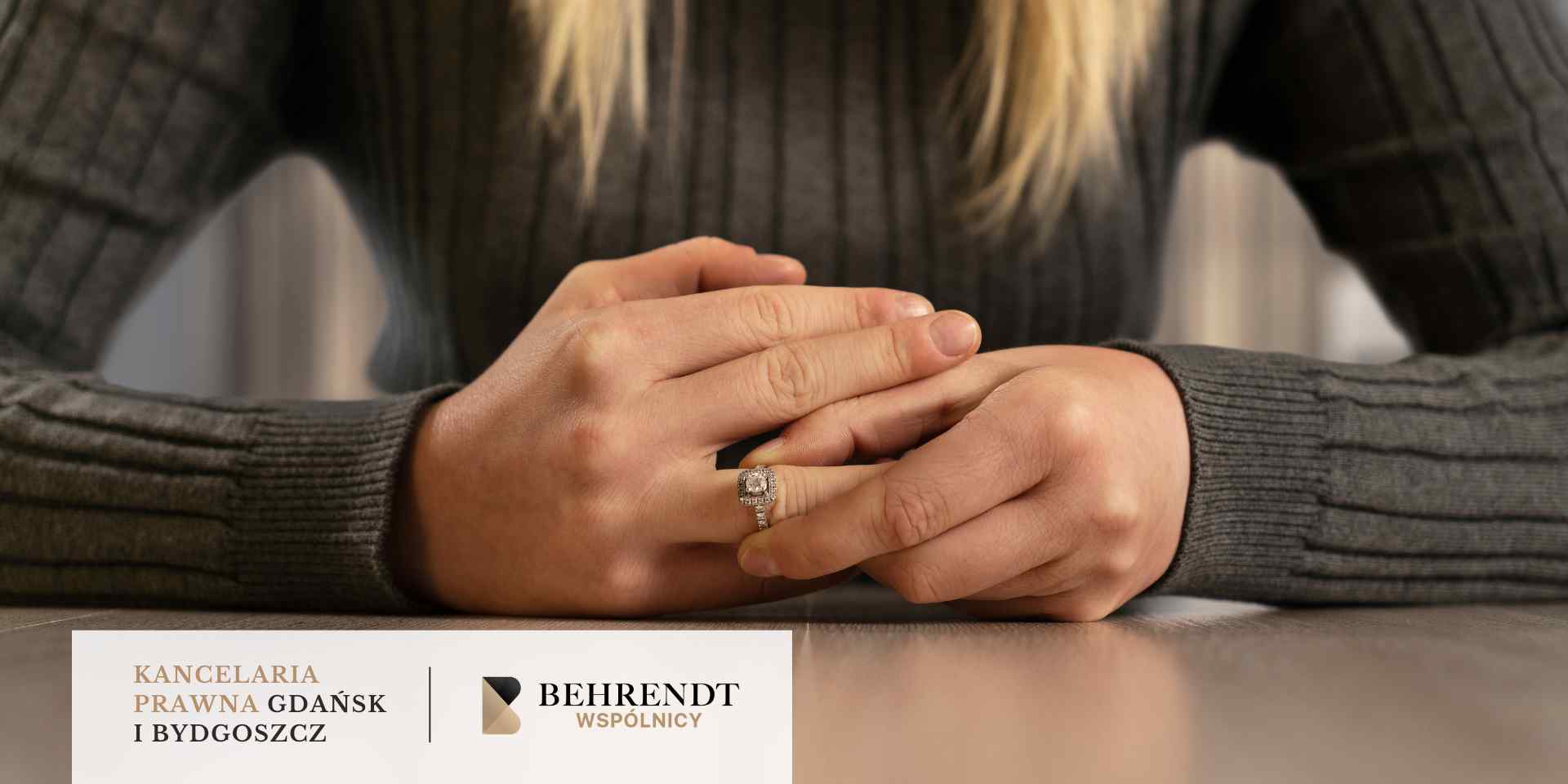 Kobieta w szarym swetrze zdejmuje z palca obrączkę na tle stołu.