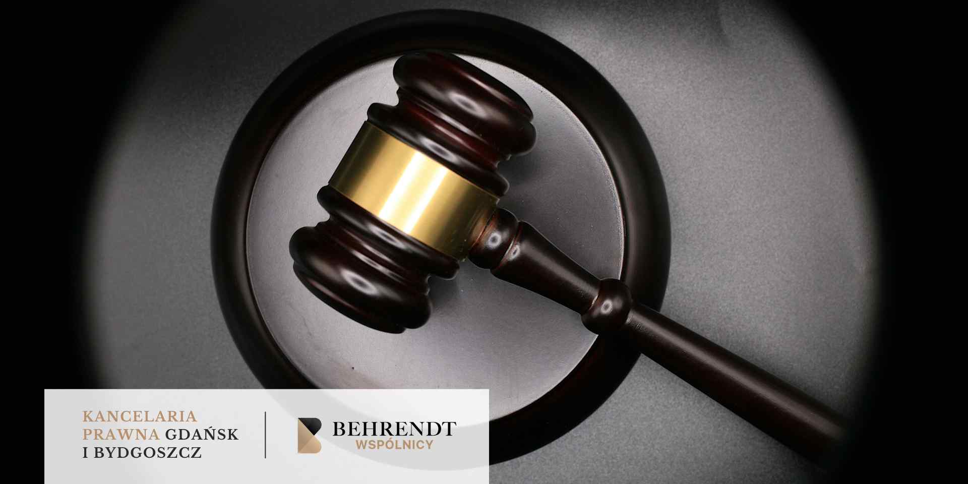 Zbliżenie na drewniany młotek sędziowski z metalową opaską, leżący na okrągłej podstawce, na ciemnym tle.