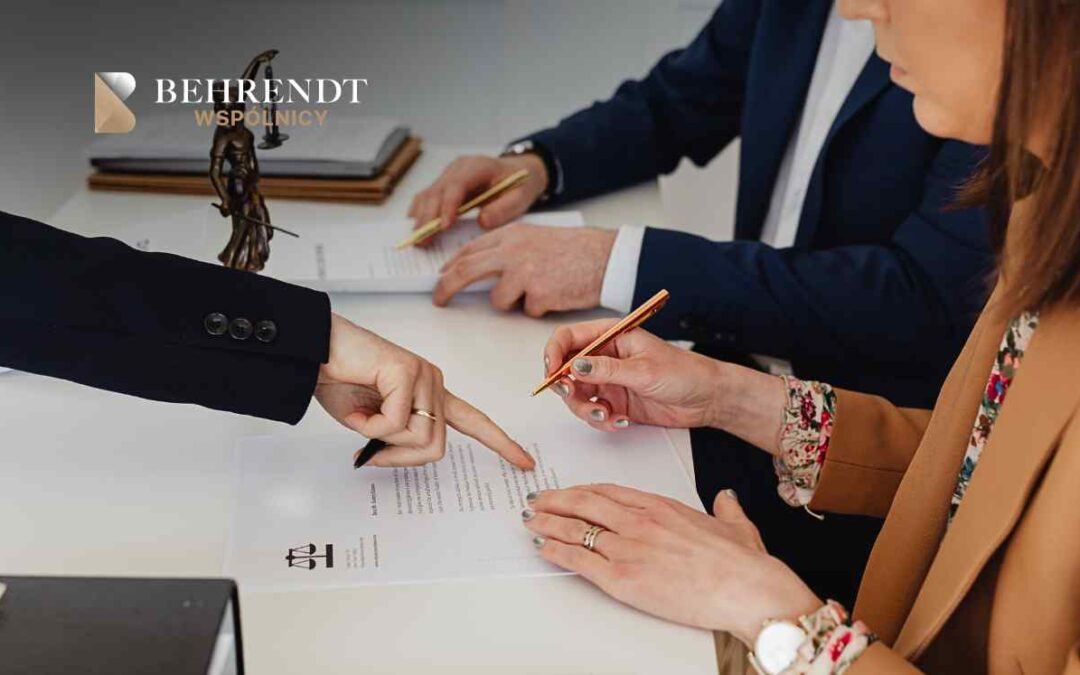 Trzy osoby w formalnym stroju siedzą przy stole i podpisują dokumenty, jedna z nich wskazuje długopisem na tekst na kartce.