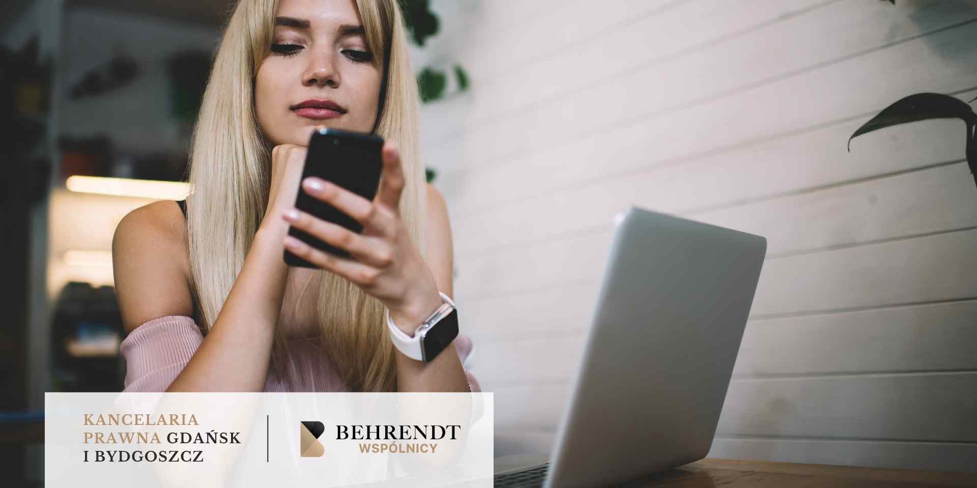 Blondynka z zamyśloną miną przegląda smartfona, siedząc przed otwartym laptopem przy drewnianym biurku.