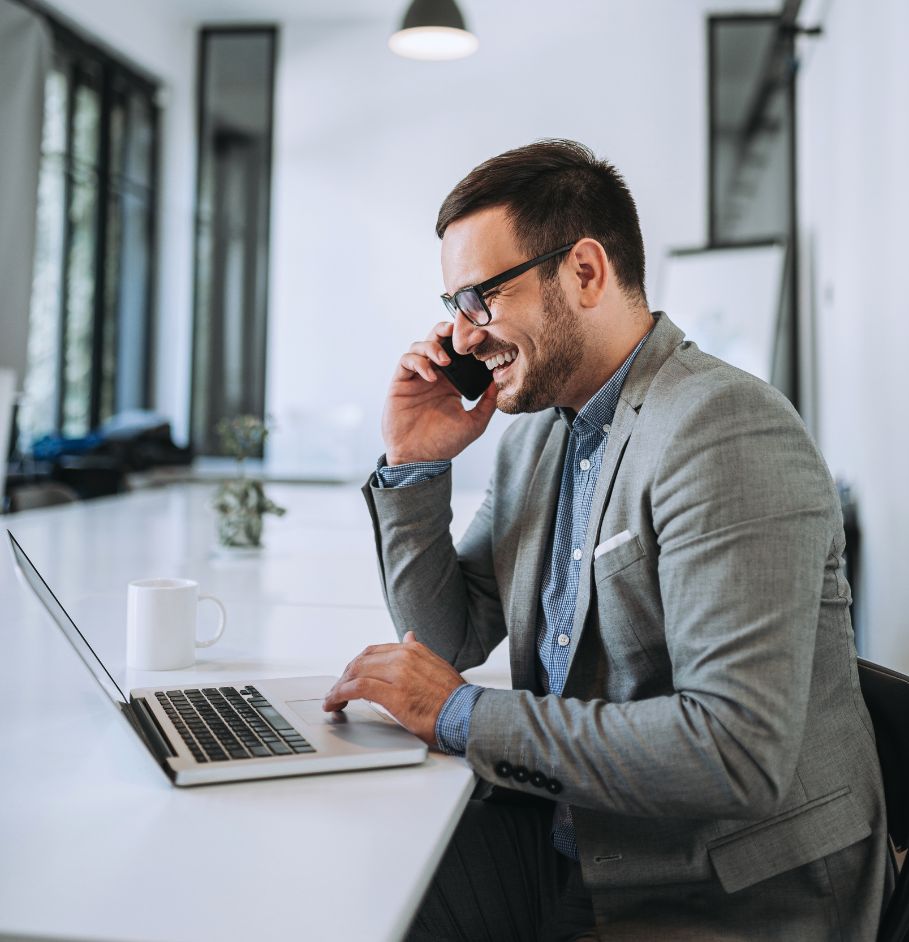 Uśmiechnięty mężczyzna w okularach i szarym garniturze rozmawia przez telefon, siedząc przed otwartym laptopem przy białym biurku.