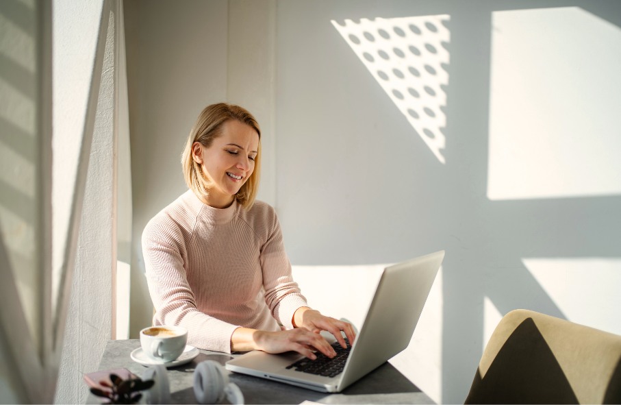 Uśmiechnięta kobieta w jasnoróżowym golfie pisze na laptopie przy stoliku, na którym stoi filiżanka kawy, w jasnym, nasłonecznionym wnętrzu.