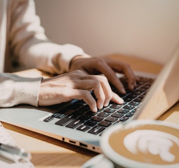 Zbliżenie na dłonie osoby w jasnym swetrze piszącej na klawiaturze laptopa, obok której na biurku stoi filiżanka kawy z wzorem latte art.