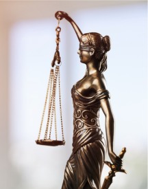 Brązowa statuetka Temidy, bogini sprawiedliwości, trzymającej wagę i miecz, przedstawiona na jasnym, rozmytym tle.