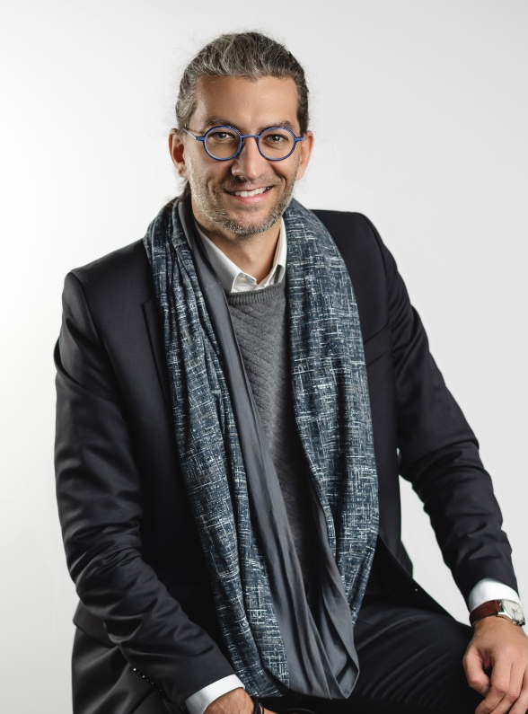 kancelaria-porady-prawne-online-trojmiasto Portret uśmiechniętego radcy prawnego Pawła Behrendta w okularach i ciemnej marynarce z ozdobnym szalem na jasnym tle.