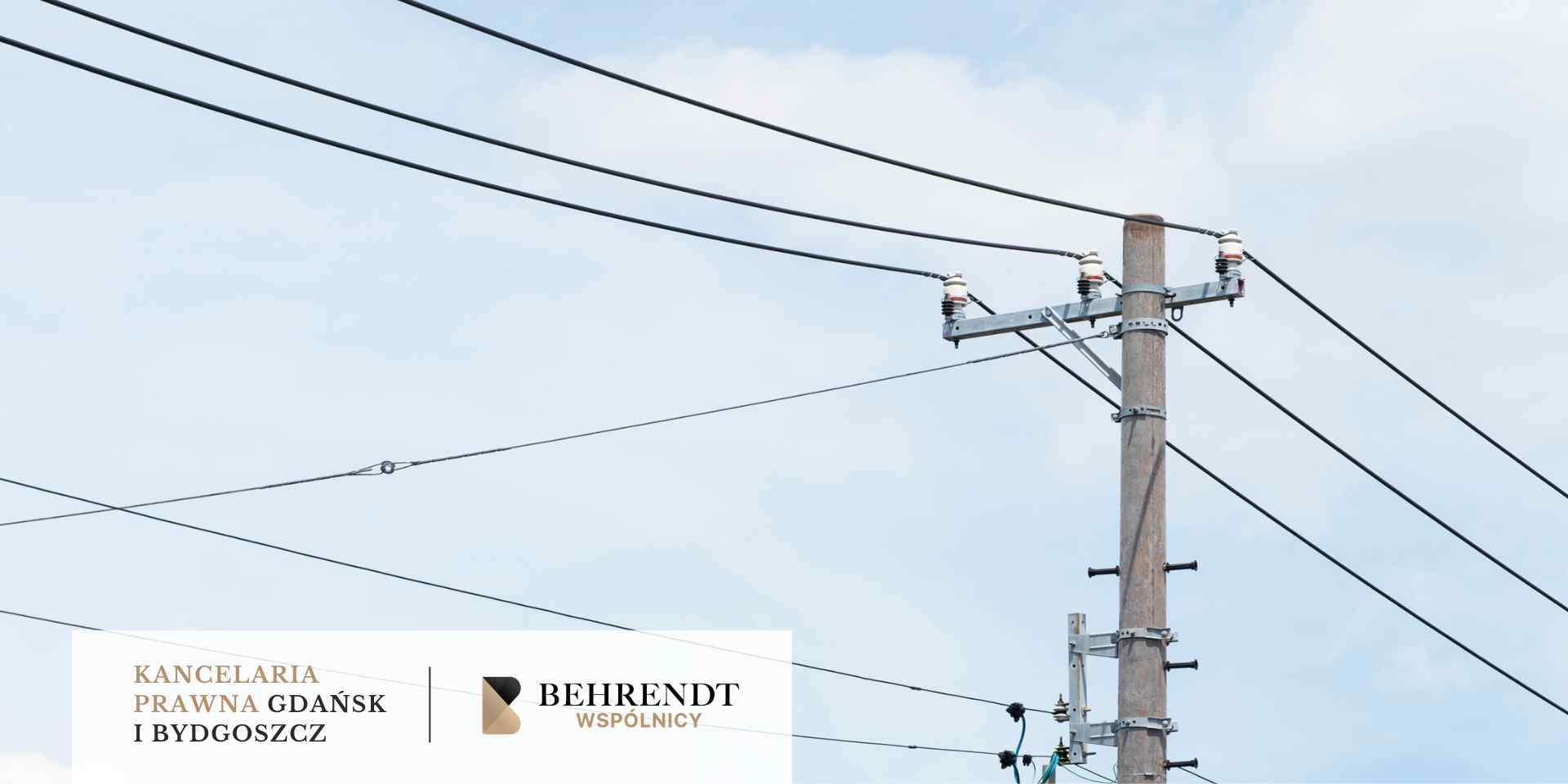 Widok na betonowy słup elektroenergetyczny z licznymi przewodami i izolatorami na tle jasnego, lekko zachmurzonego nieba.