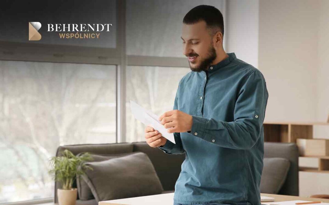 Młody mężczyzna z brodą, ubrany w niebieską koszulę, stoi w jasnym biurze i z uśmiechem przegląda dokumenty wyjęte z białej koperty. W tle widoczne są duże okna oraz nowoczesne wnętrze kancelarii.