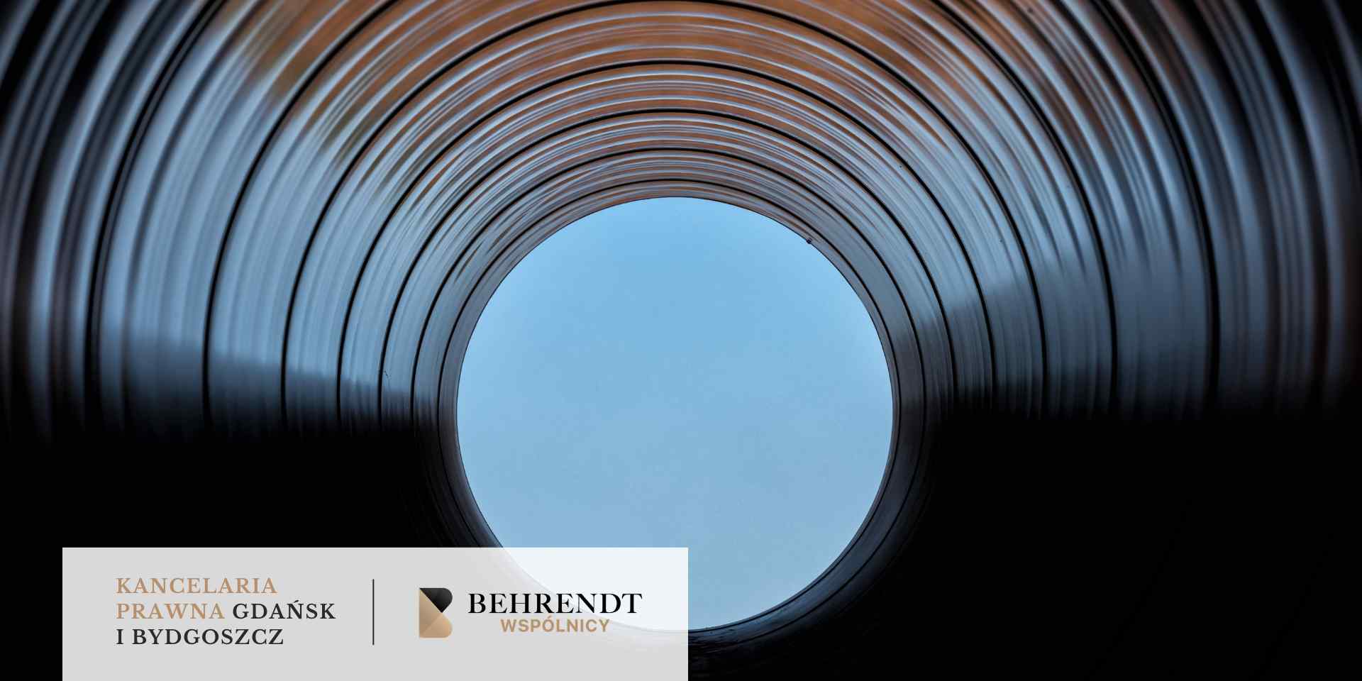 Perspektywiczne ujęcie z wnętrza stalowej rury o dużym przekroju, ukazujące koncentryczne kręgi metalowych żłobień prowadzące do jasnego, błękitnego otworu (nieba) na końcu.