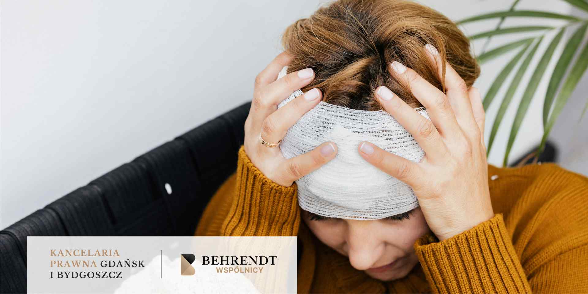 Kobieta w żółtym swetrze siedzi ze spuszczoną głową, trzymając się dłońmi za boki głowy owiniętej szerokim, białym opatrunkiem medycznym.