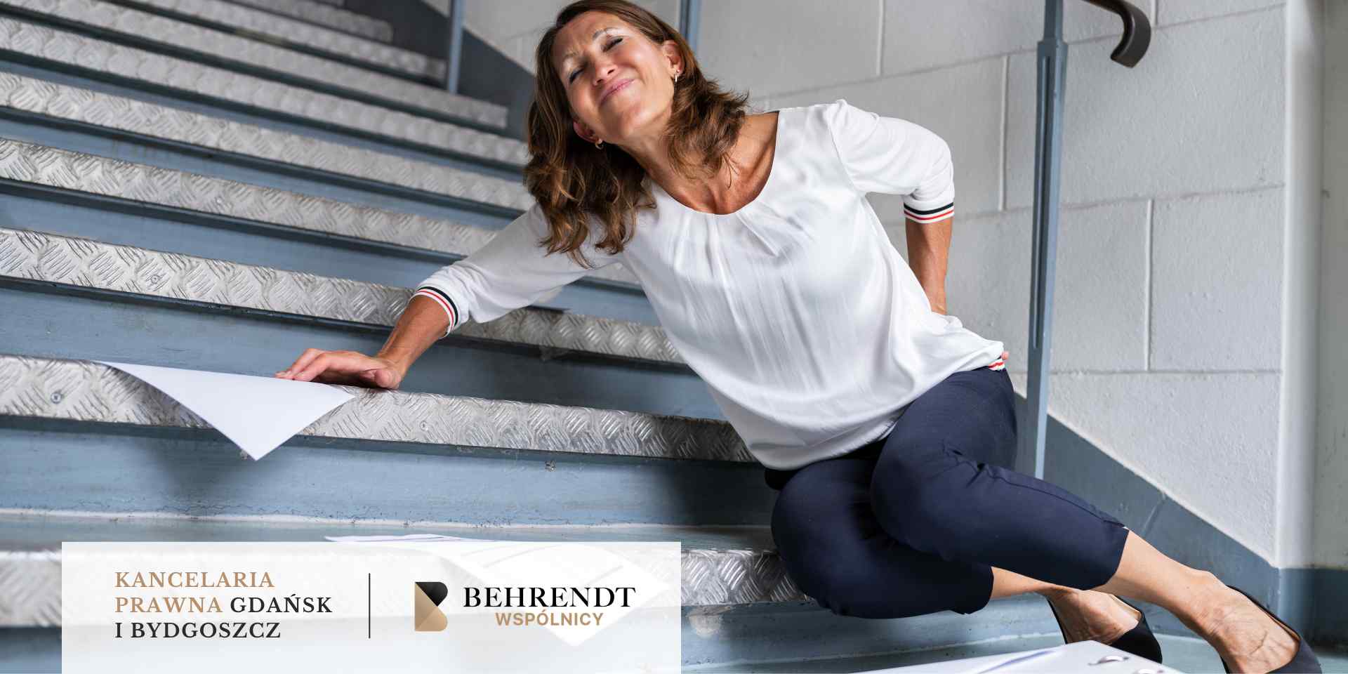 Kobieta siedząca na metalowych schodach z grymasem bólu, trzymająca się za dolną część pleców po upadku; obok niej leżą rozrzucone dokumenty.