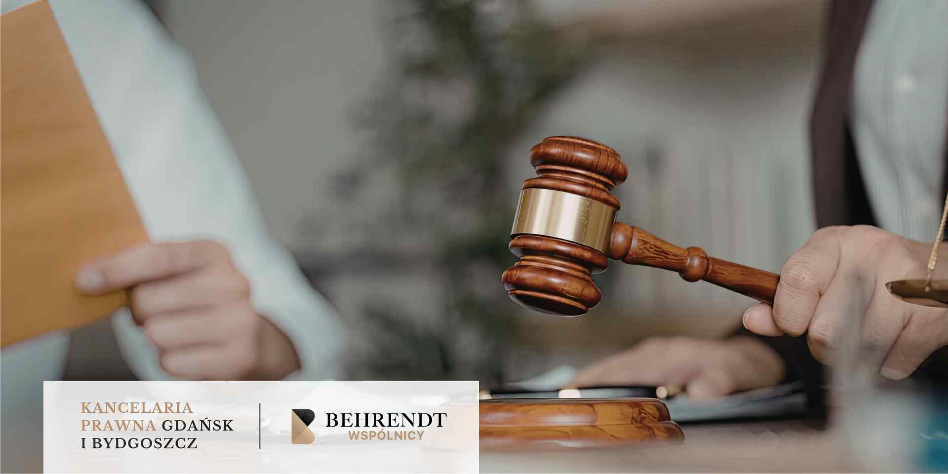 Zbliżenie na dłoń sędziego uderzającego drewnianym młotkiem w podstawkę, obok której stoi waga szalkowa; w tle widoczna jest postać trzymająca dokumenty.