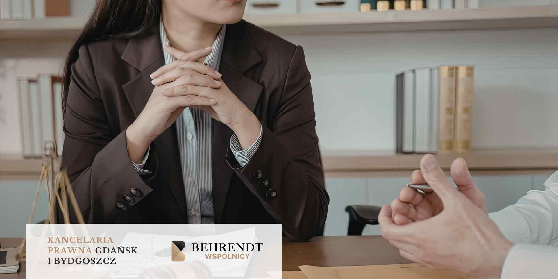 Kobieta w brązowej marynarce siedzi przy biurku z zaplecionymi dłońmi, uważnie słuchając osoby siedzącej naprzeciwko, której dłoń z długopisem widoczna jest w prawym dolnym rogu.