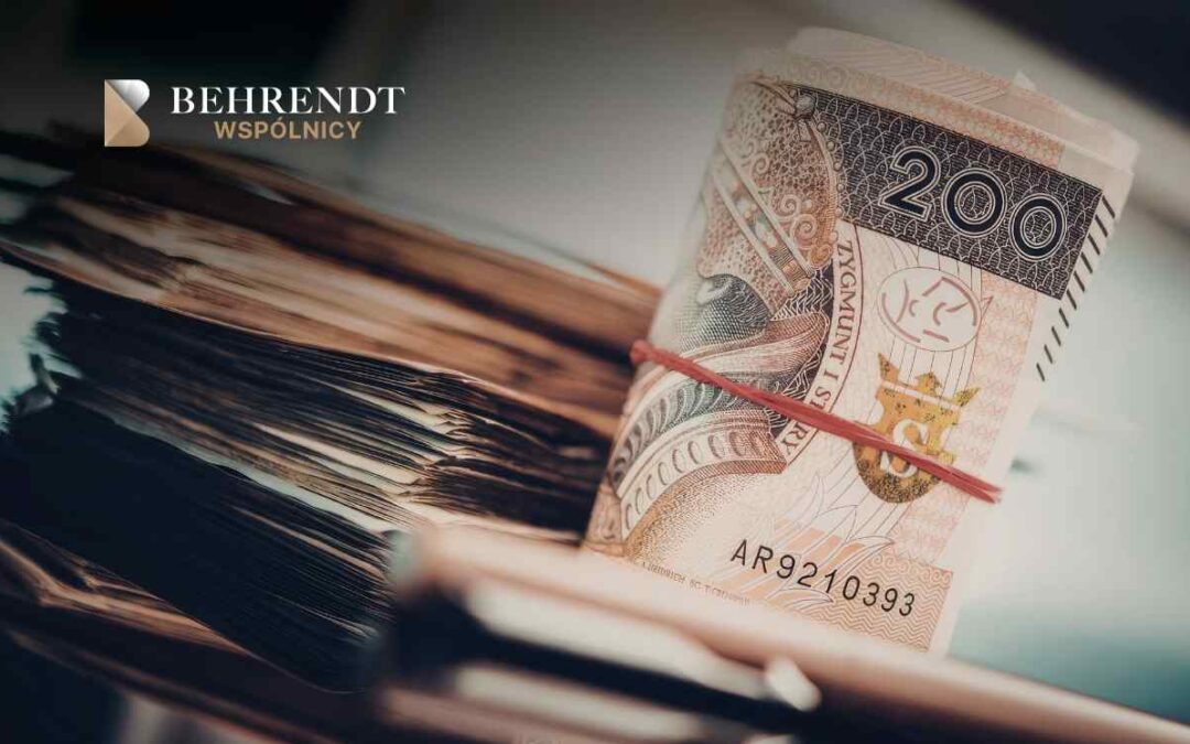 Zbliżenie na gruby plik dokumentów (prawdopodobnie akt prawnych) leżących na biurku, obok których stoi rulon banknotów o nominale 200 złotych polskich (PLN), spięty czerwoną gumką recepturką. W lewym górnym rogu znajduje się logotyp Kancelarii Behrendt Wspólnicy.