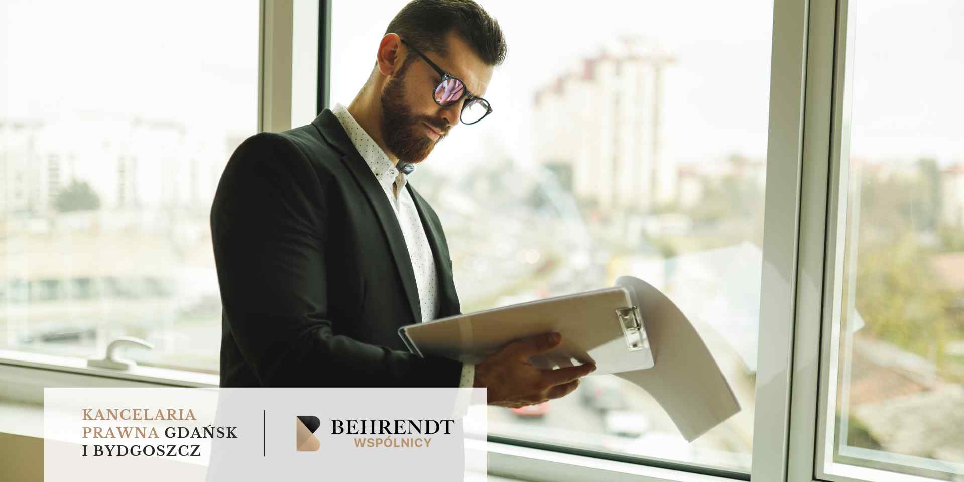 Mężczyzna w ciemnej marynarce i okularach stoi przy dużym oknie biurowym, uważnie analizując dokumenty umieszczone w białym folderze. W tle widoczna jest panorama miasta.