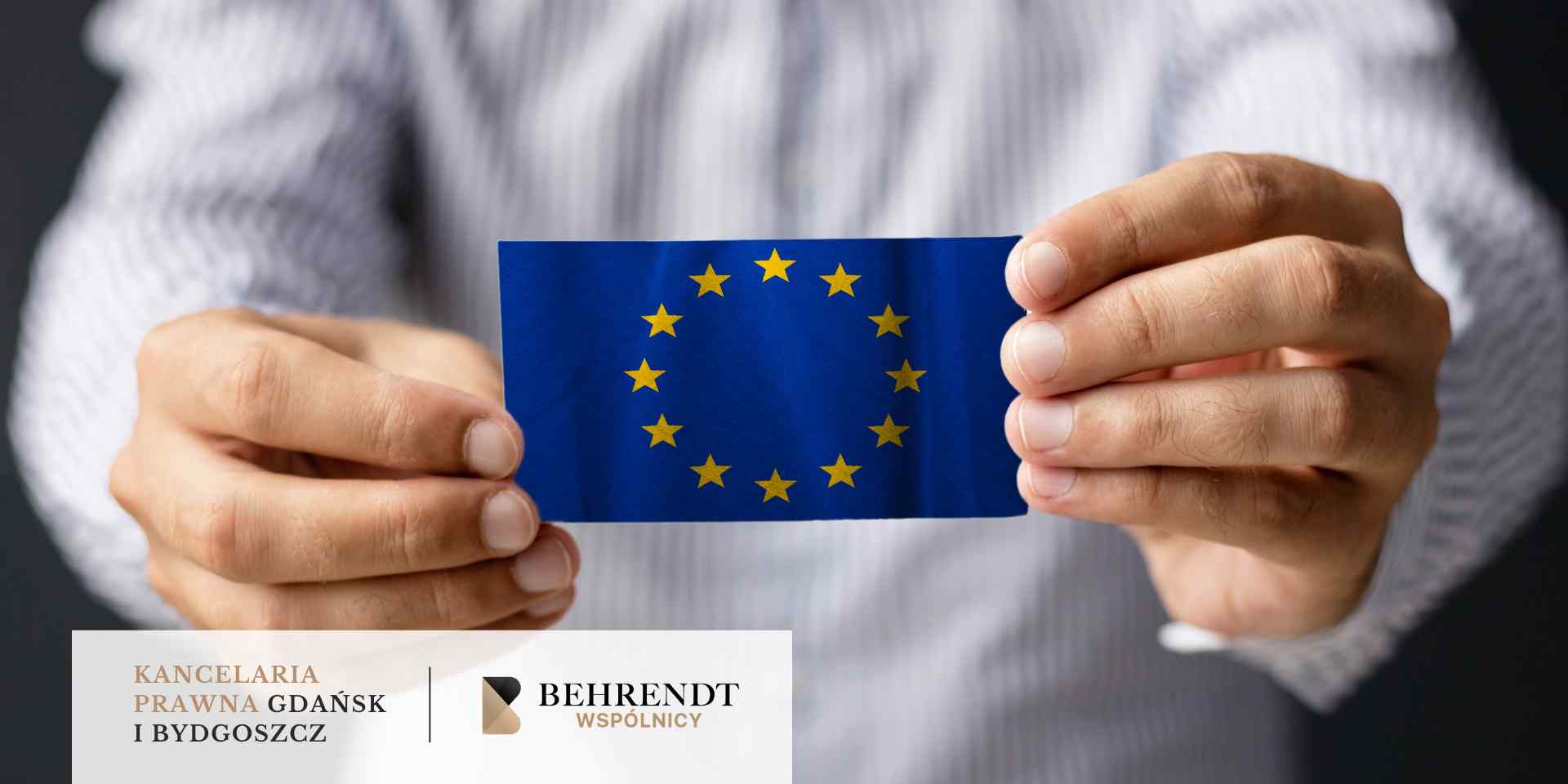 Zbliżenie na dłonie osoby w jasnej koszuli, trzymającej oburącz małą flagę Unii Europejskiej.