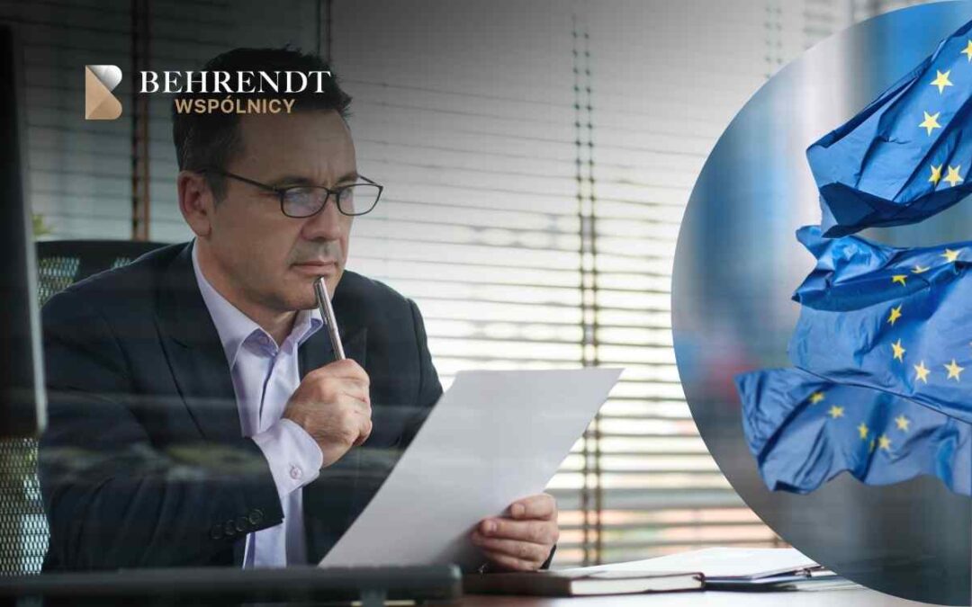 Prawnik w ciemnej marynarce siedzi przy biurku w biurze, trzymając długopis przy ustach w geście głębokiego zastanowienia podczas lektury dokumentu. Po prawej stronie wkomponowano grafikę flagi Unii Europejskiej, a w lewym górnym rogu widnieje logotyp Kancelarii Behrendt Wspólnicy.