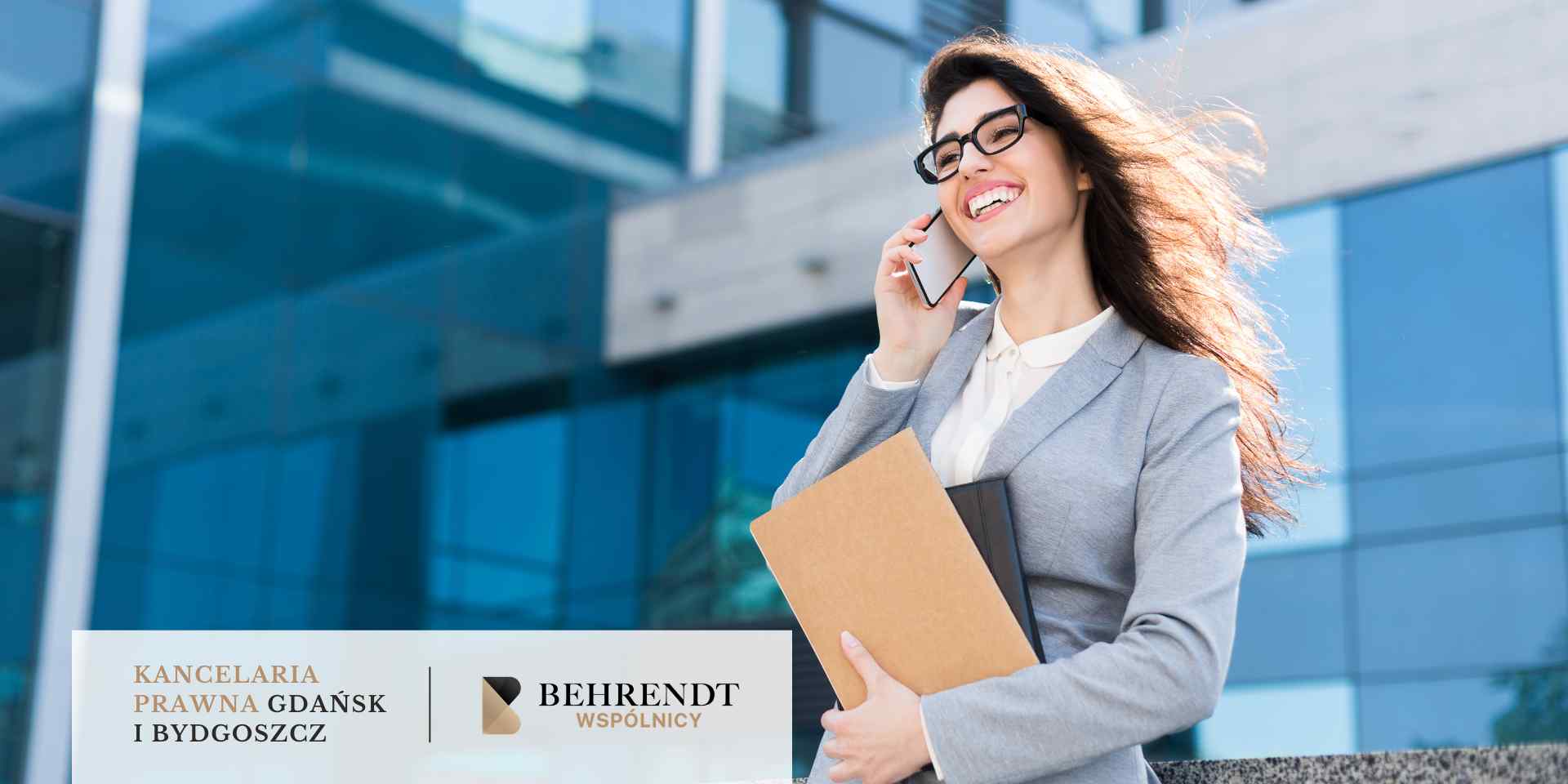 Uśmiechnięta prawniczka w okularach i szarym żakiecie rozmawia przez telefon, trzymając teczkę z dokumentami na tle nowoczesnego biurowca. W dolnej części grafiki znajduje się biały pasek z logotypem Kancelarii Behrendt Wspólnicy z Gdańska i Bydgoszczy.