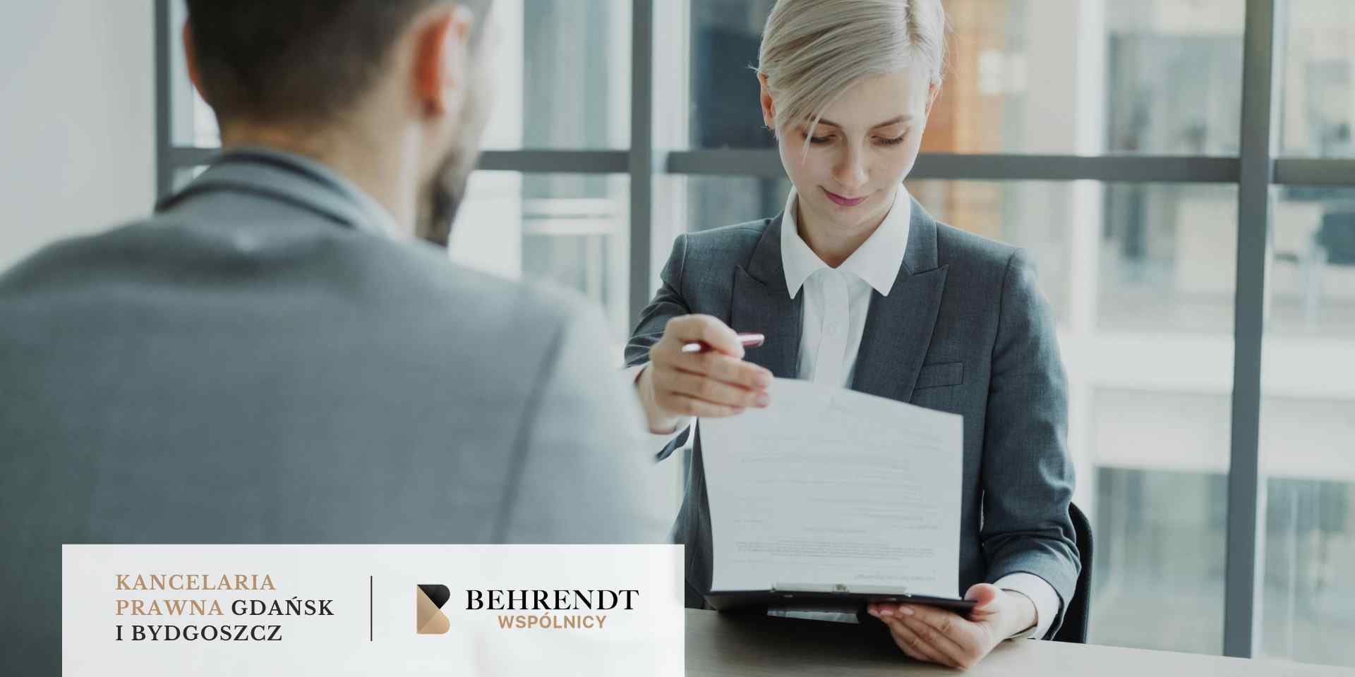 Kobieta w ciemnoszarej marynarce (prawniczka) siedzi przy biurku naprzeciwko klienta (widocznego od tyłu). Z lekkim uśmiechem i profesjonalnym skupieniem prezentuje klientowi dokument umieszczony w czarnym folderze, wskazując długopisem na konkretny zapis.