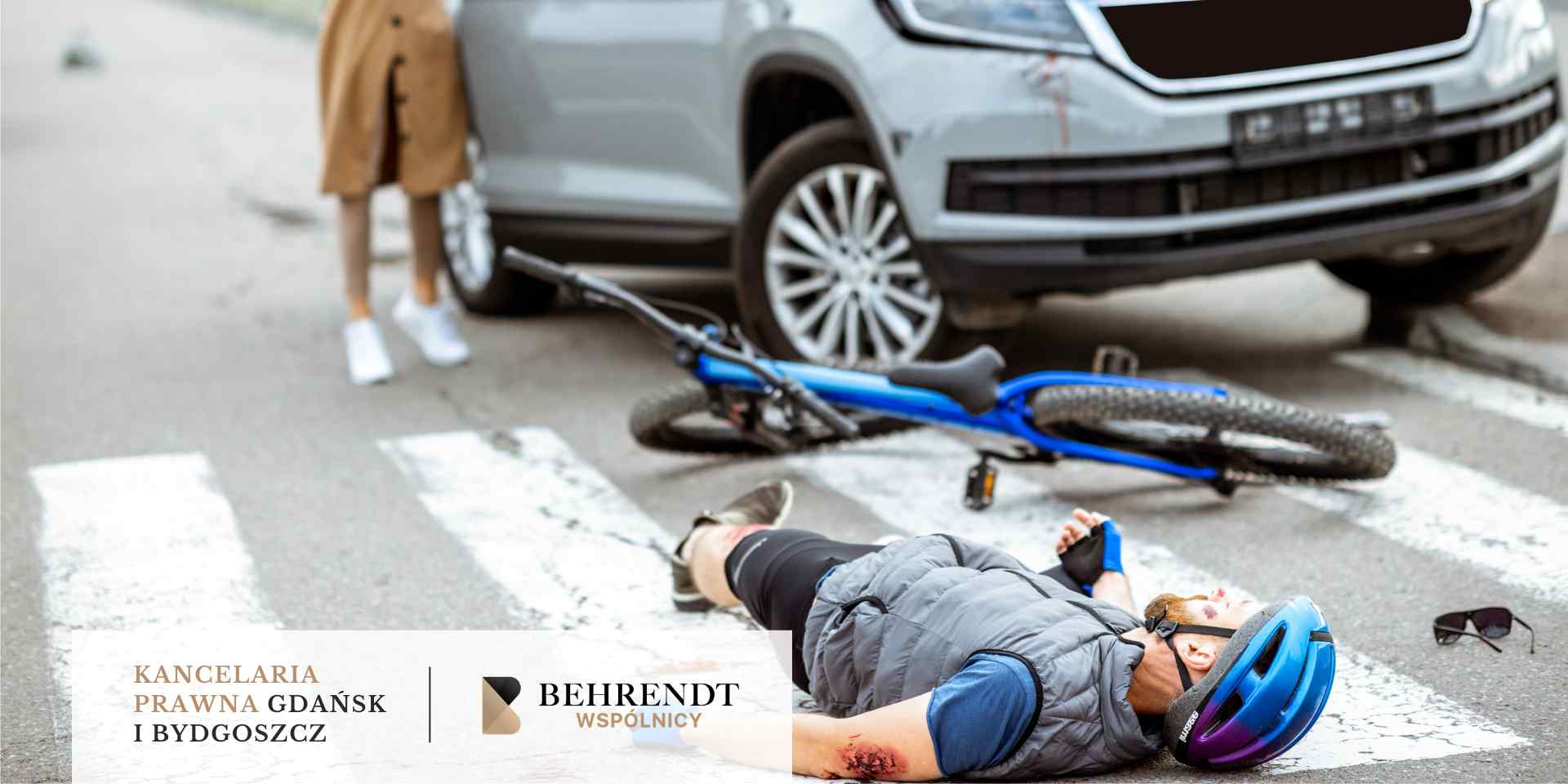 Mężczyzna w kasku rowerowym leży na przejściu dla pieszych po potrąceniu przez samochód; obok leży przewrócony niebieski rower, a w tle widać przód srebrnego auta.