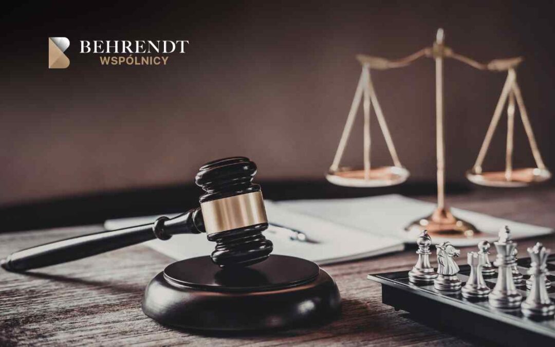 Na drewnianym blacie znajdują się symbole wymiaru sprawiedliwości oraz strategii: czarny młotek sędziowski na podstawce, waga szalkowa w tle oraz rozstawione na szachownicy srebrne figury szachowe.