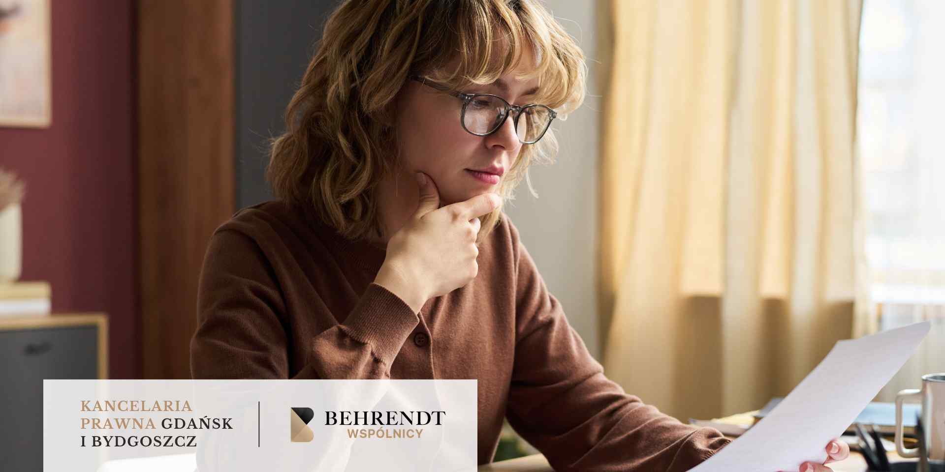 Młody mężczyzna z brodą, ubrany w niebieską koszulę, stoi w jasnym biurze i z uśmiechem przegląda dokumenty wyjęte z białej koperty.