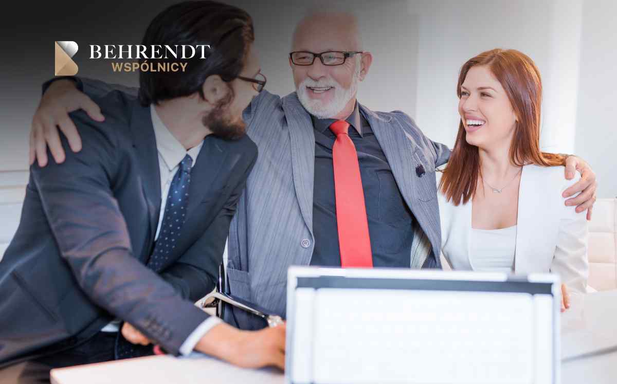 Three business professionals smiling and leaning together at a desk, Behrendt WSPÓLNICY logo visible in the top left corner.
