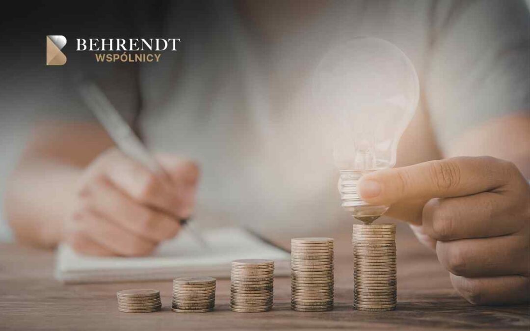 Stacks of coins increasing in height on a wooden desk; a hand places a lightbulb-shaped coin on the tallest stack, symbolizing investment growth, with Behrendt Wspólnicy branding in the corner.