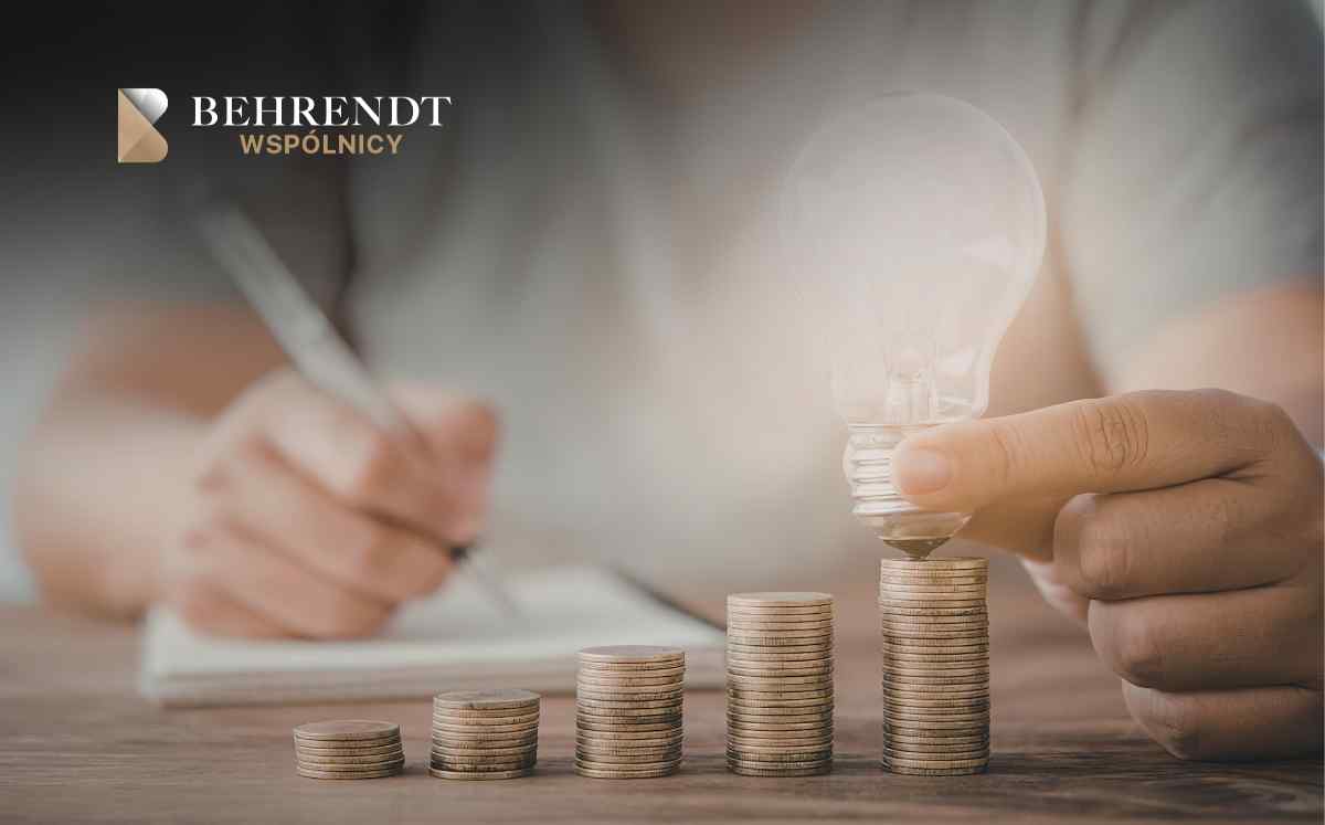 Stacks of coins increasing in height on a wooden desk; a hand places a lightbulb-shaped coin on the tallest stack, symbolizing investment growth, with Behrendt Wspólnicy branding in the corner.