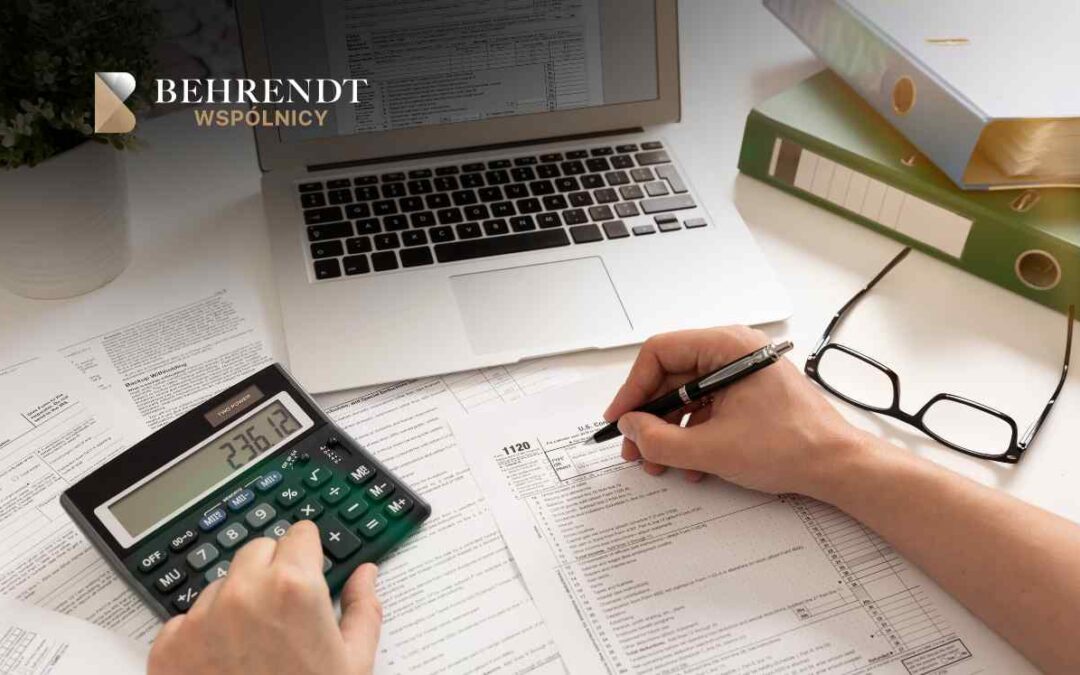 Accountant's desk with a hand writing on tax forms, a calculator, laptop, glasses, and stacked binders nearby.