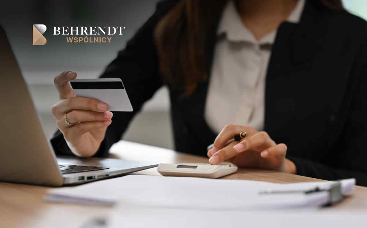 Businesswoman using a laptop and calculator at a desk, holding a card; logo 'Behrendt Wspólnicy' visible in top left corner.