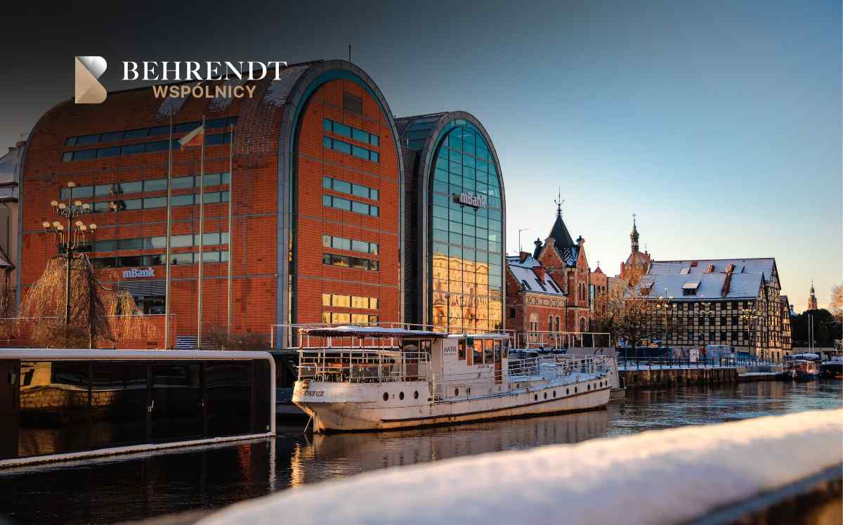 Logo 'BEHRENDT WSPÓLNICY' on a brick and glass riverside building with a white boat docked by the water.