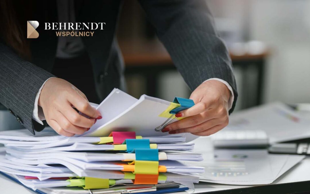 Businessperson in a suit organizing a stack of papers with colorful sticky notes on a desk, Behrendt Wspólnicy logo in the top-left corner.