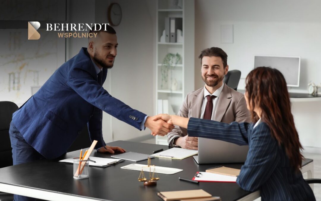 Handshake across a desk between a man in a blue suit and a woman in a striped blazer, with a smiling colleague nearby in a modern офис setting. Logo Behrendt Wspólnicy in the corner.