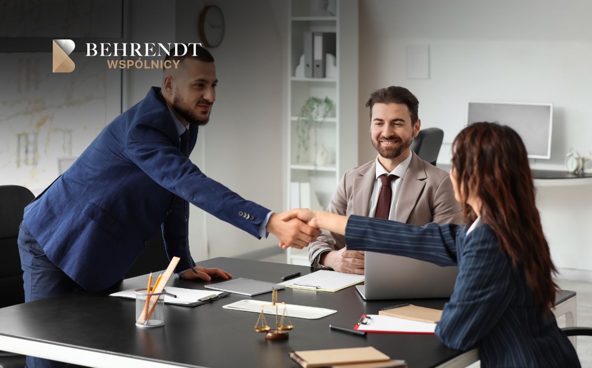 Handshake across a desk between a man in a blue suit and a woman in a striped blazer, with a smiling colleague nearby in a modern офис setting. Logo Behrendt Wspólnicy in the corner.