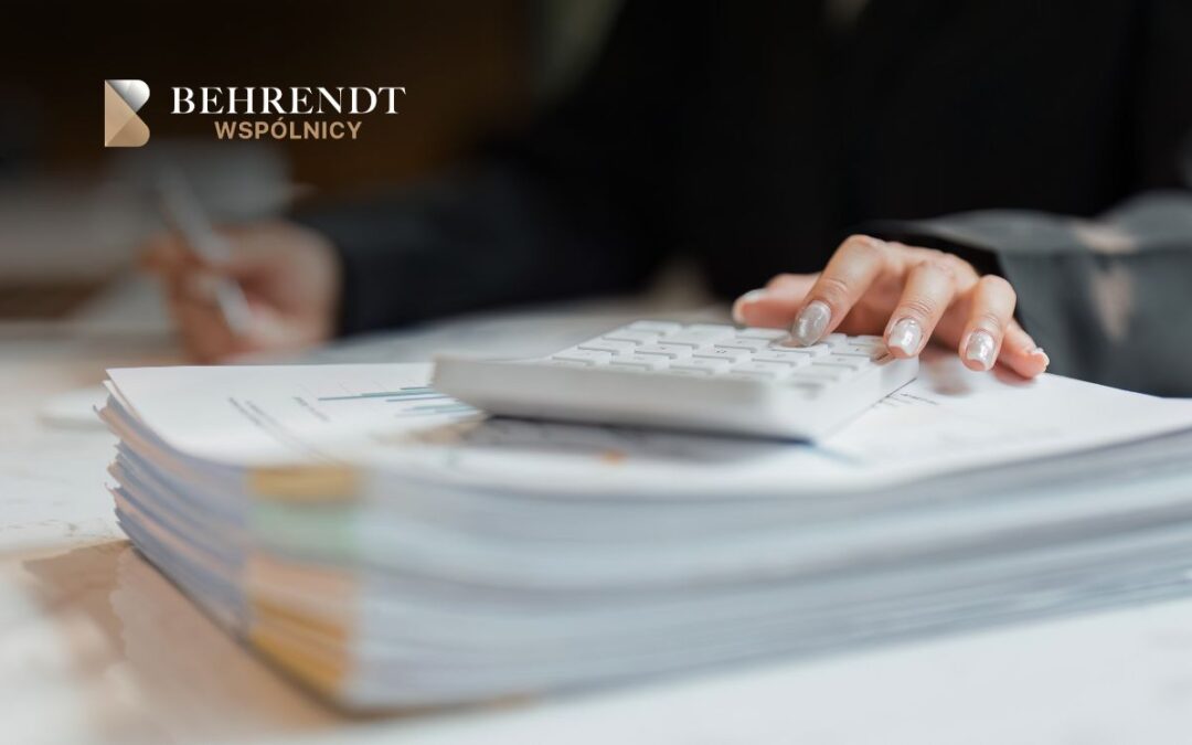 Hand on a calculator beside a stack of papers on a desk, with the Behrendt Wspólnicy logo in the top-left corner.