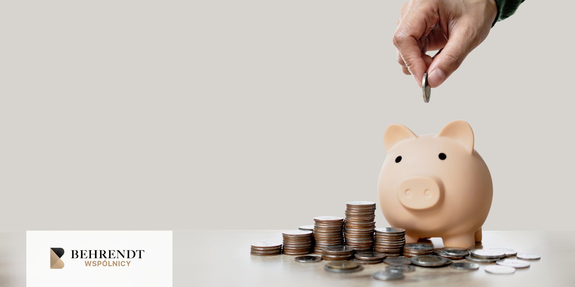 Zbliżenie na rękę wrzucającą od góry monetę do świnki skarbonki. Obok niej, na biurku znajdują się monety